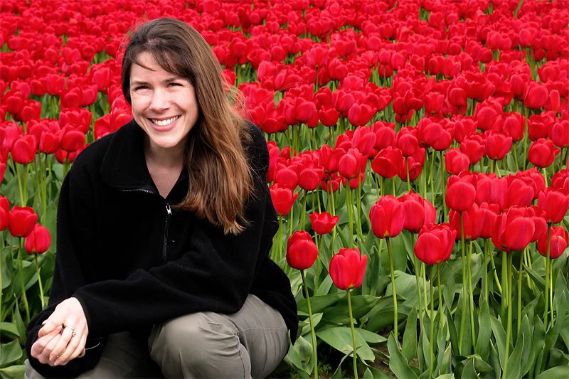 A woman with long brown hair is smiling while sitting among vibrant red tulips. She appears relaxed and happy amidst the colorful flower field.