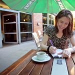 Depeche Mode A woman smiles while jotting notes in a notebook outside, accompanied by her coffee. She sits under a green umbrella on a wooden patio table.
