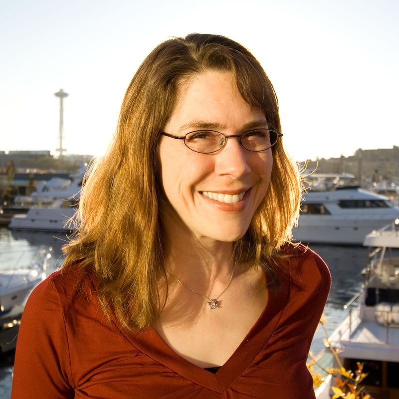 Kristie, wearing glasses and a red top, smiles warmly in front of a marina with the iconic Seattle Space Needle visible in the distance.