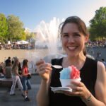 The Rohdenburgs Kristie smiles while holding a red and blue shaved ice treat. A bustling fountain scene is in the background.