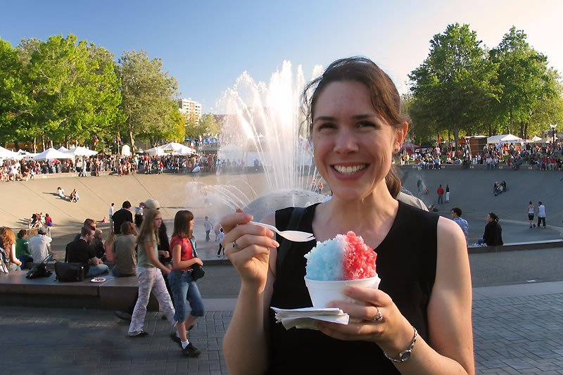 Kristie smiles while holding a red and blue shaved ice treat. A bustling fountain scene is in the background.