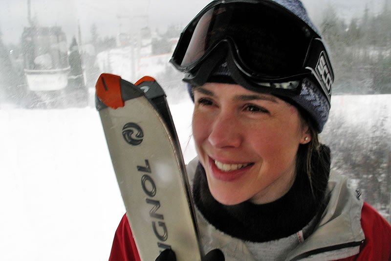 Kristie wearing ski goggles and a beanie, smiling while holding skis. A snowy mountain landscape is visible in the background.