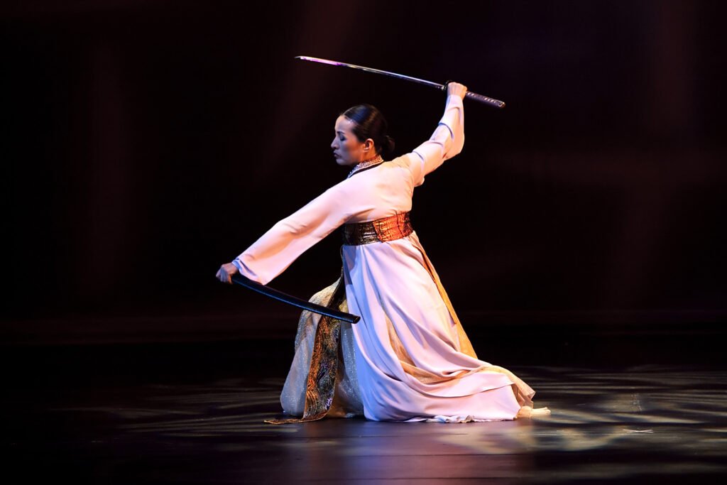 A dancer performs a graceful sword routine on stage wearing traditional white attire.