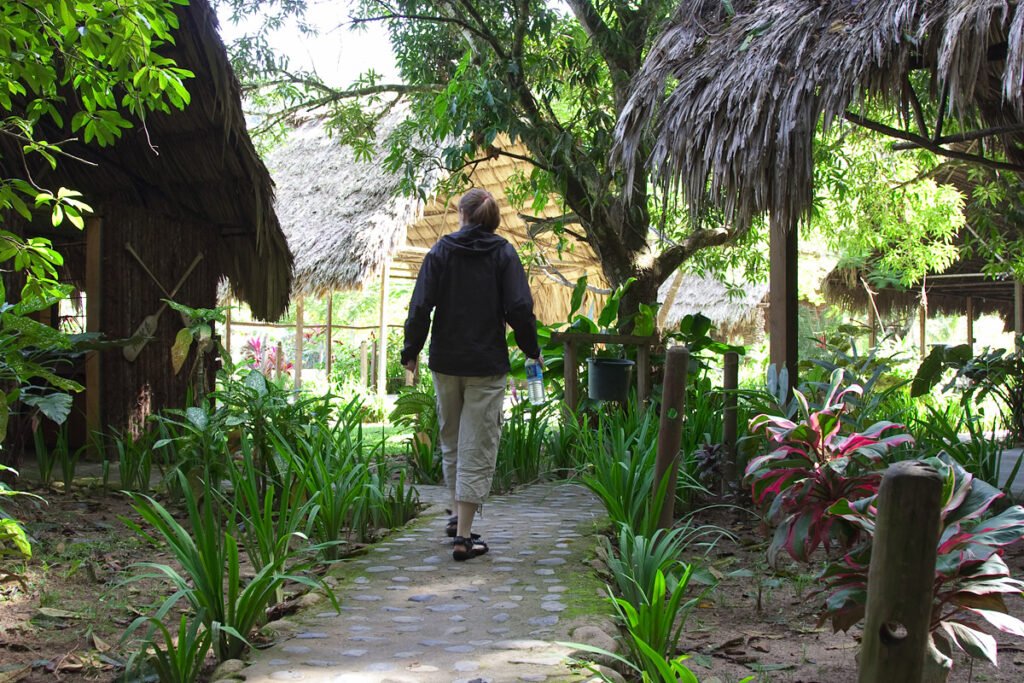 Kristie walks down a lush garden pathway lined with vibrant plants and natural thatched structures.
