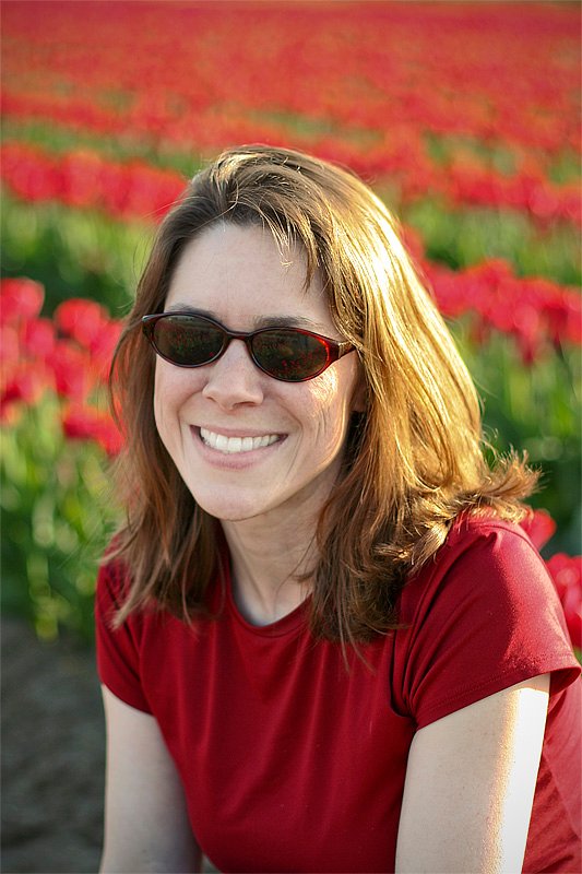 Kristie wearing sunglasses and a red shirt smiles in front of vibrant red tulips.