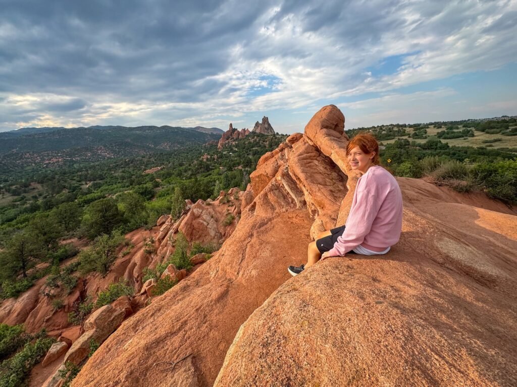 Kristie sits on a large red rock formation, gazing out over a vast landscape filled with greenery and distant hills under a cloudy sky.