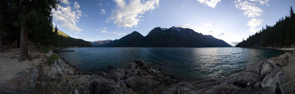 A panoramic view of a tranquil lake surrounded by dense forests and majestic mountains under a clear blue sky.