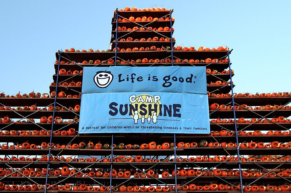 Massive metal racks stacked high with orange pumpkins under a clear blue sky feature a large light blue banner. The banner displays the Life is Good smiley face logo and the text for Camp Sunshine.