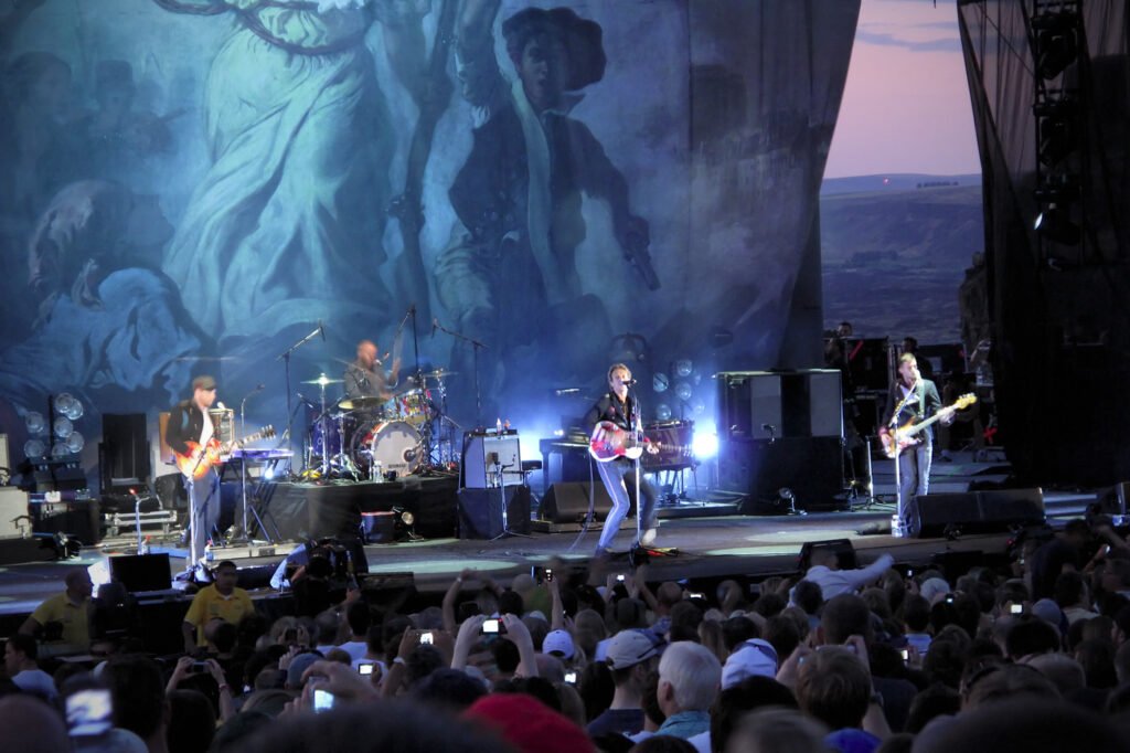 A live concert stage performance featuring a band playing guitars and surrounded by an energetic audience recording the show.