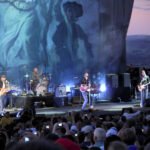 Sand Point A live concert stage performance featuring a band playing guitars and surrounded by an energetic audience recording the show.