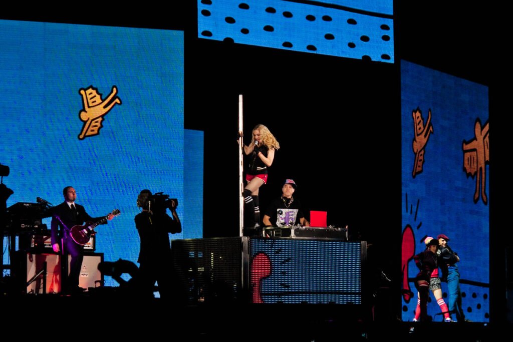 A dynamic live music performance featuring a guitarist and a pole dancer with vibrant blue and orange graphics in the background.
