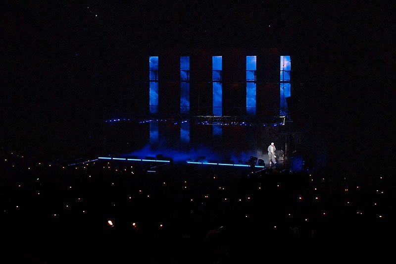 A performer illuminated by dramatic blue lights on stage with an enthusiastic crowd holding up lights.