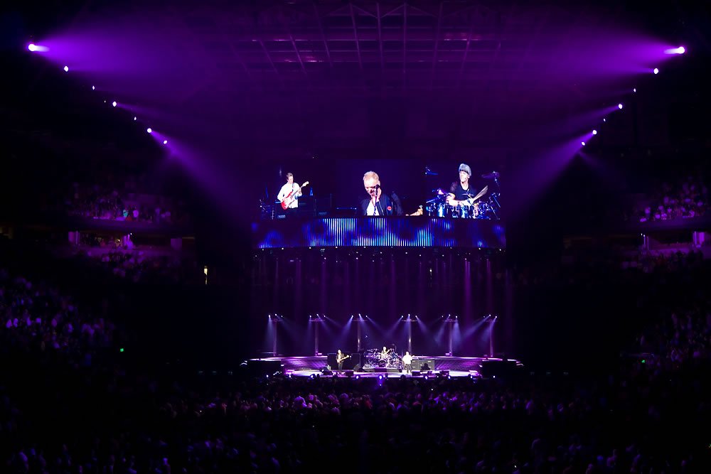 A band performing on stage under vibrant purple lighting with an engaged audience in the background.