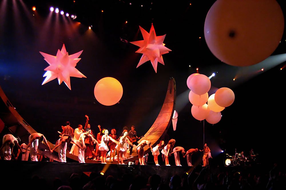 A live concert stage with performers and colorful decorations. The scene features large spherical and star-shaped ornaments.