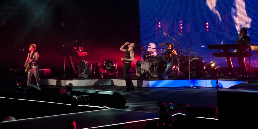 A live concert scene featuring a band performing on stage under vibrant red and blue lighting. Musicians play various instruments including guitars, drums, and synthesizers.