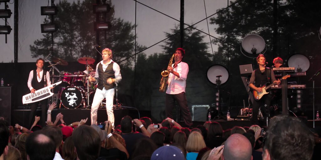 A rock band performs on stage, engaging an enthusiastic crowd. The singer wears a white outfit, while other musicians play guitars, keyboards, and a saxophone.