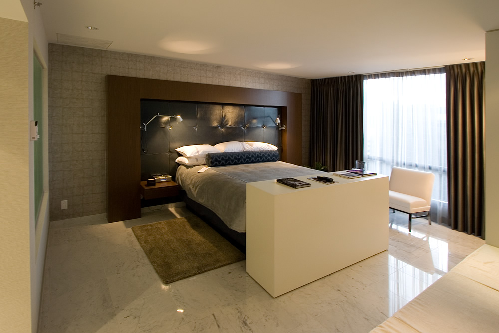 A modern hotel room featuring a sleek bed, a white rectangular bench, and large windows with curtains.