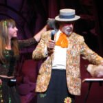 A magician in a colorful outfit is performing a trick with a live rat. He is dressed in a vibrant jacket and a hat, holding a microphone, while a young assistant stands beside him.