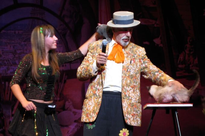 A magician in a colorful outfit is performing a trick with a live rat. He is dressed in a vibrant jacket and a hat, holding a microphone, while a young assistant stands beside him.