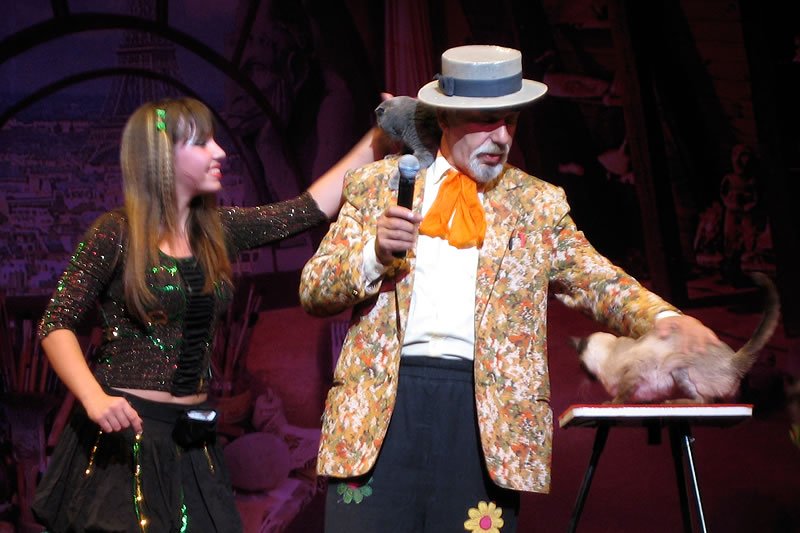 A magician in a colorful outfit is performing a trick with a live rat. He is dressed in a vibrant jacket and a hat, holding a microphone, while a young assistant stands beside him.