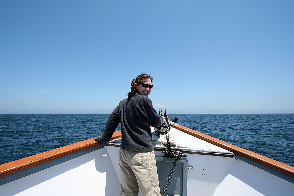 A man with sunglasses and a ponytail stands at the bow of a sailboat. The open sea meets a clear blue sky.