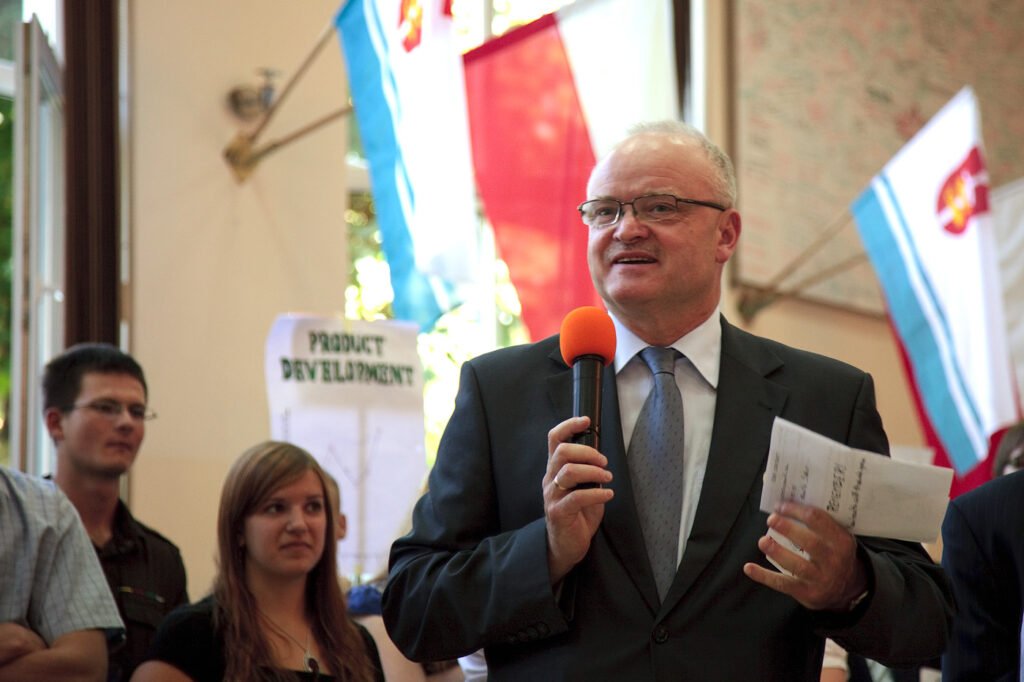 A man in a suit stands speaking into a microphone, with flags and an attentive audience in the background.