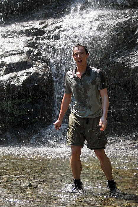 A man stands under a cascading waterfall, water droplets around him. He is dressed casually and appears to be enjoying the refreshing experience.