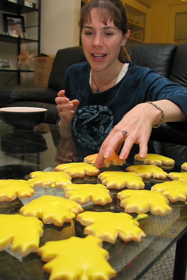 A woman is enjoying maple leaf-shaped cookies with yellow icing on a glass table. She is seated comfortably at home, surrounded by cozy decor.