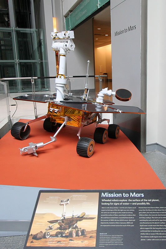 A detailed model of a Mars rover on display in an exhibit, with informational text and images below it. The rover is situated on an orange platform representing the Martian surface.