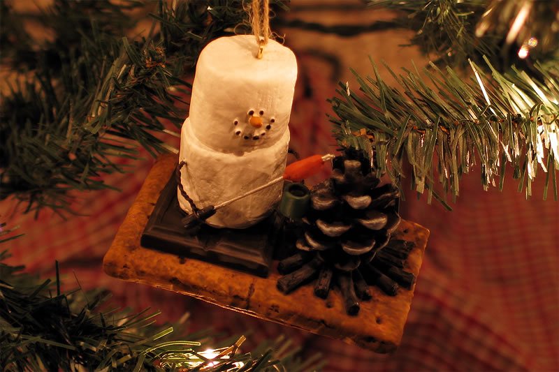 A cozy Marshmallow Snowman Christmas ornament hangs from a branch, capturing holiday charm. The miniature snowman is crafted from stacked marshmallows with a small pinecone for a base.