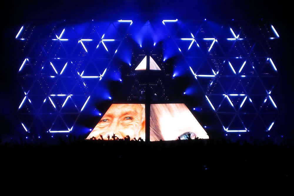 A striking blue light display with geometric patterns illuminates the stage, creating an immersive atmosphere for concertgoers. The vibrant lights contrast against the dark surroundings, captivating the audience below.