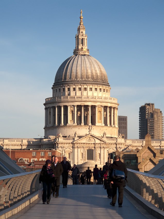 A scenic view of St. Paul's Cathedral in London as seen from the Millennium Bridge. Pedestrians are walking along the bridge, with the sun casting a warm glow on the cathedral's dome.
