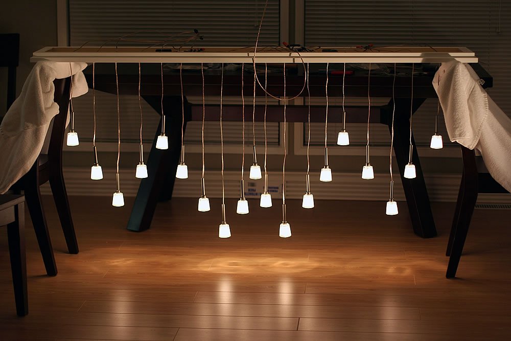 A modern chandelier with multiple hanging bulbs suspended from a wooden frame, casting warm light onto a wooden floor.