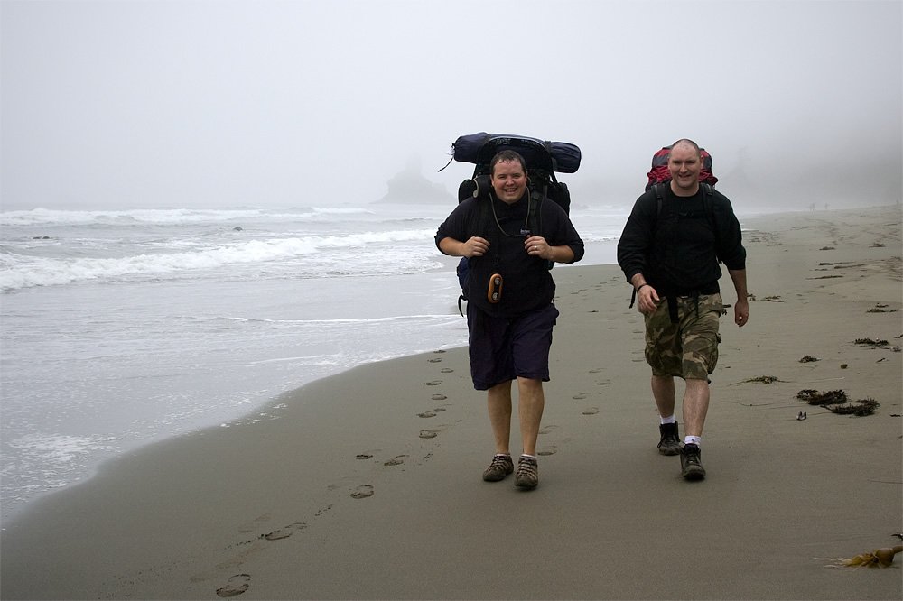 Two hikers, John and Kristie, trek along a foggy Pacific Northwest beach, carrying large backpacks and leaving footprints in the sand.