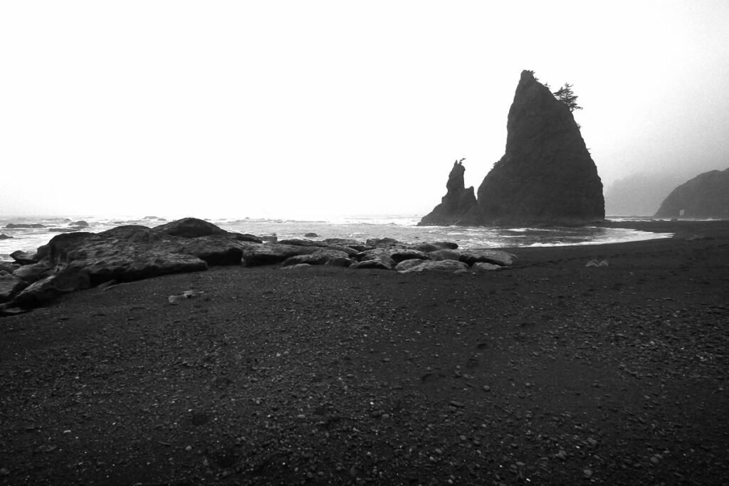 A black and white photo of a misty black sand beach with large rock formations in the distance.