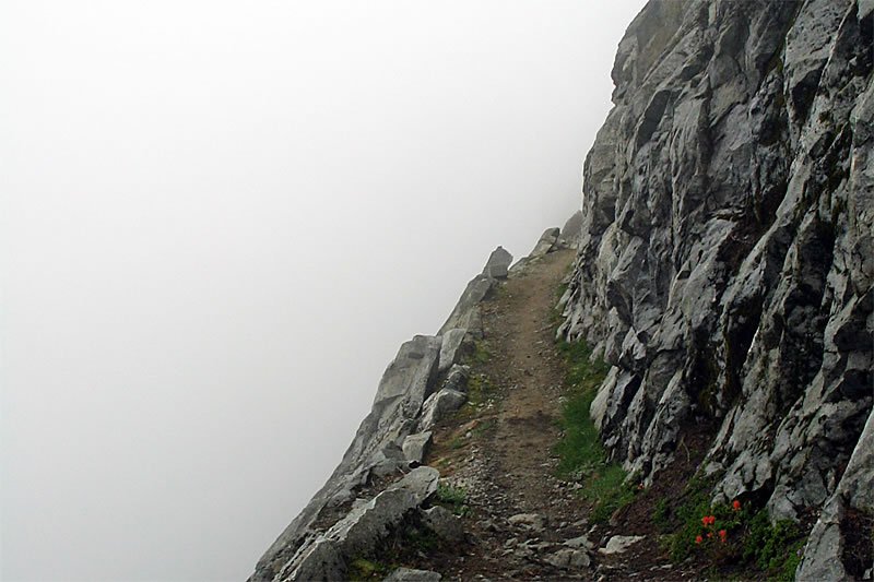 A narrow trail winds through rocky terrain, partially obscured by dense fog in a Pacific Northwest mountain setting.