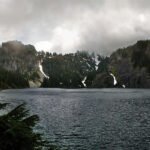 The Crystal Method A serene lake framed by dense evergreen trees, with mist rising from the surrounding hills under a cloudy sky.