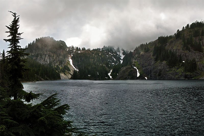 A serene lake framed by dense evergreen trees, with mist rising from the surrounding hills under a cloudy sky.