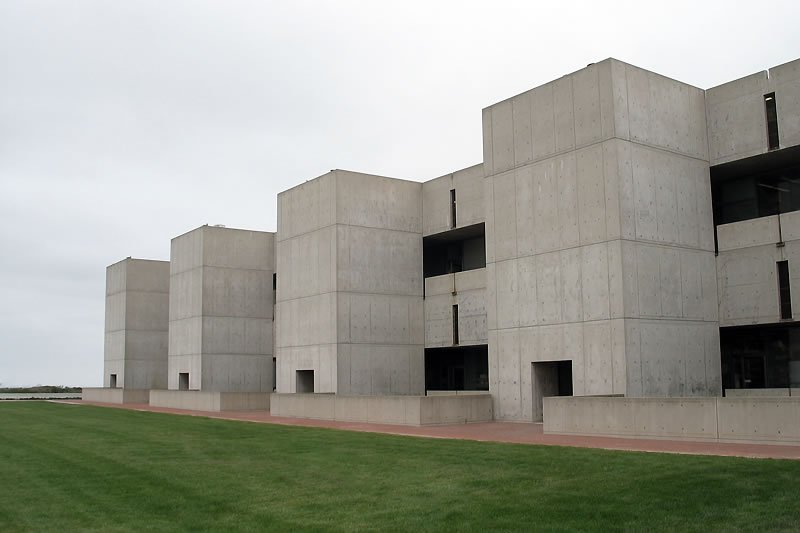 A row of modern concrete buildings adjacent to a lush green lawn under an overcast sky.