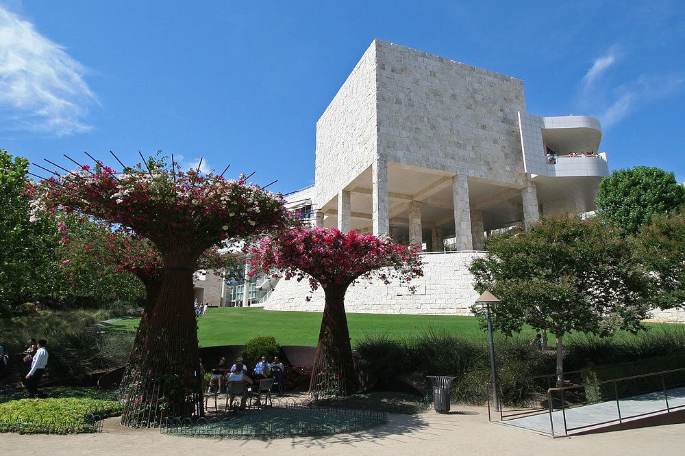 A contemporary building with white walls and large windows, surrounded by lush greenery and vibrant purple-flowered trees. People are seated on benches enjoying the serene environment under a clear blue sky.