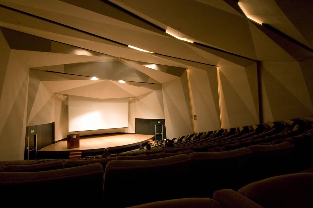An empty auditorium with rows of dark seats facing a brightly lit stage under modern lighting fixtures.