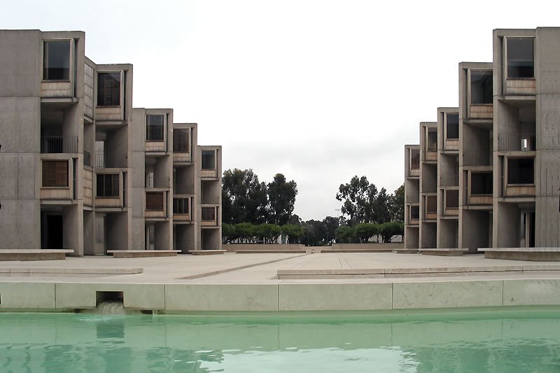 A row of modern, multi-story buildings with boxy designs reflecting in a serene pool of water.