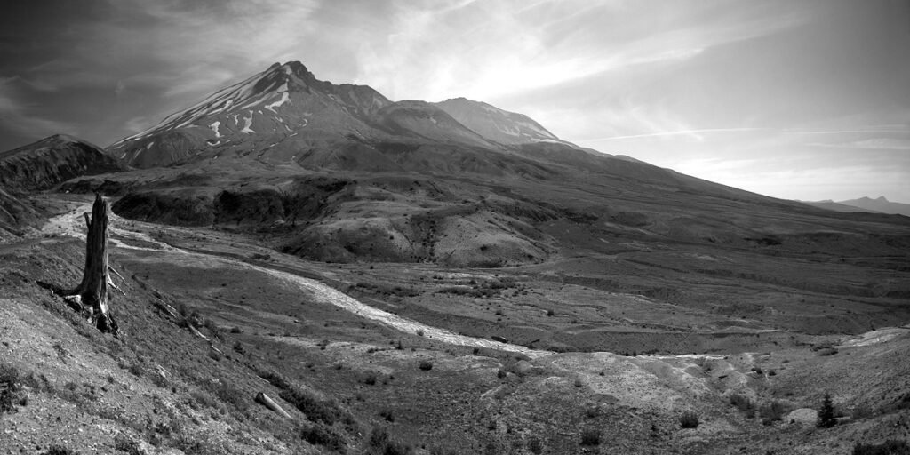 A lone hiker stands on a mountain trail, looking towards the majestic, snow-capped peak of Mount Rainier under a cloudy sky.