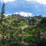 Three Fingers A tranquil mountain lake flanked by dense pine forests. The calm water mirrors the surrounding greenery and blue sky.