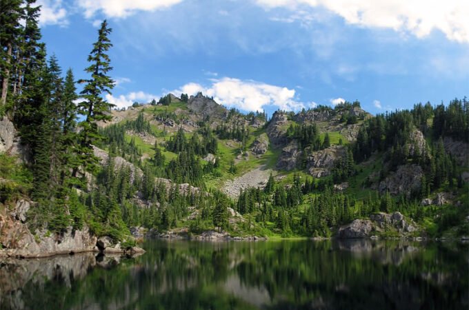 A tranquil mountain lake flanked by dense pine forests. The calm water mirrors the surrounding greenery and blue sky.