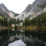 The Rohdenburgs A calm alpine lake mirrors towering granite peaks and evergreen trees under an overcast sky. Lush forest slopes descend to the water’s edge, creating a tranquil natural scene.