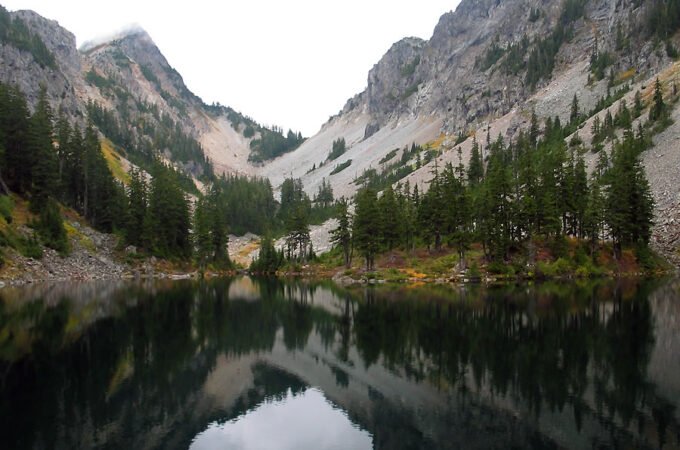 A calm alpine lake mirrors towering granite peaks and evergreen trees under an overcast sky. Lush forest slopes descend to the water’s edge, creating a tranquil natural scene.