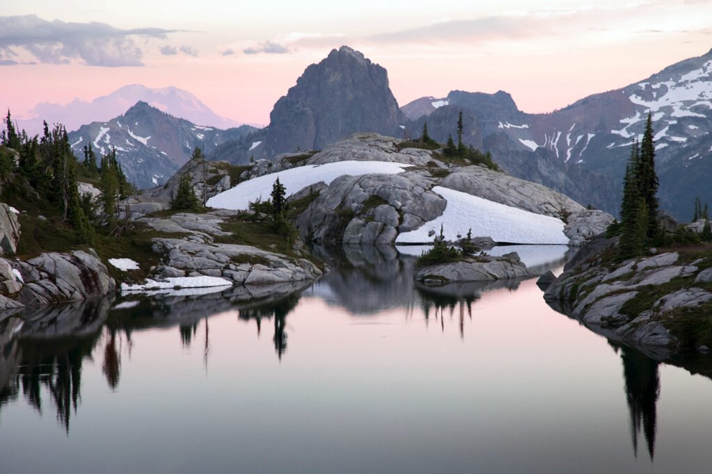A tranquil mountain lake surrounded by snow-covered rocks and trees, reflecting the soft pink sky.