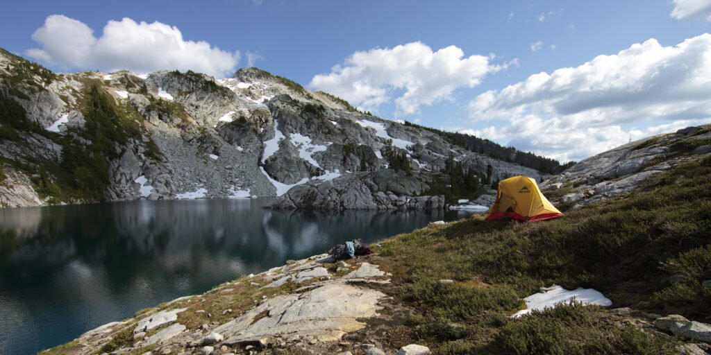 A tranquil campsite next to a serene mountain lake, with a yellow tent set up on rocky terrain under a bright blue sky, surrounded by lush greenery and distant mountain peaks.