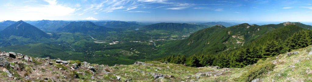 A panoramic view from the top of a mountain showing vast green valleys and distant peaks under a clear blue sky.
